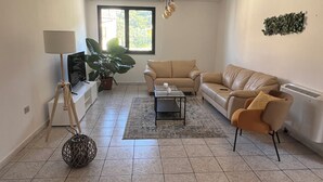 Uma sala de estar ampla com um sofá bege, uma mesa de centro e uma TV sobre um suporte. Há uma janela grande que oferece luz natural e vista para o verde lá fora.