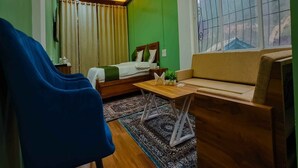 A room with a bed, a sofa, a blue chair, a small table, and a large window with a view. - Mudita Hotel (West Kameng)