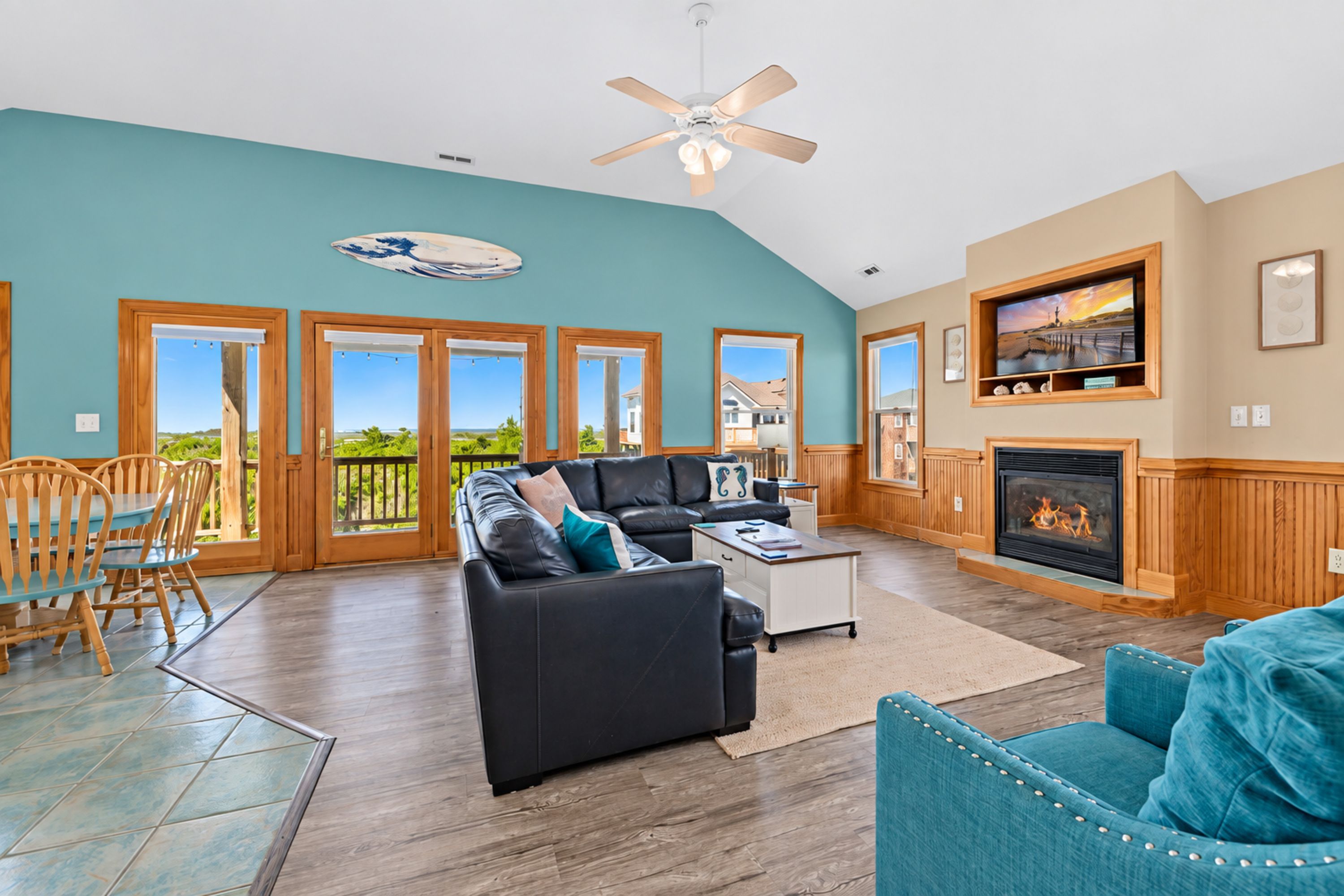 A spacious living area with a black leather sofa, a coffee table, and a fireplace. There are wooden doors leading to a balcony with a view of the outdoors.