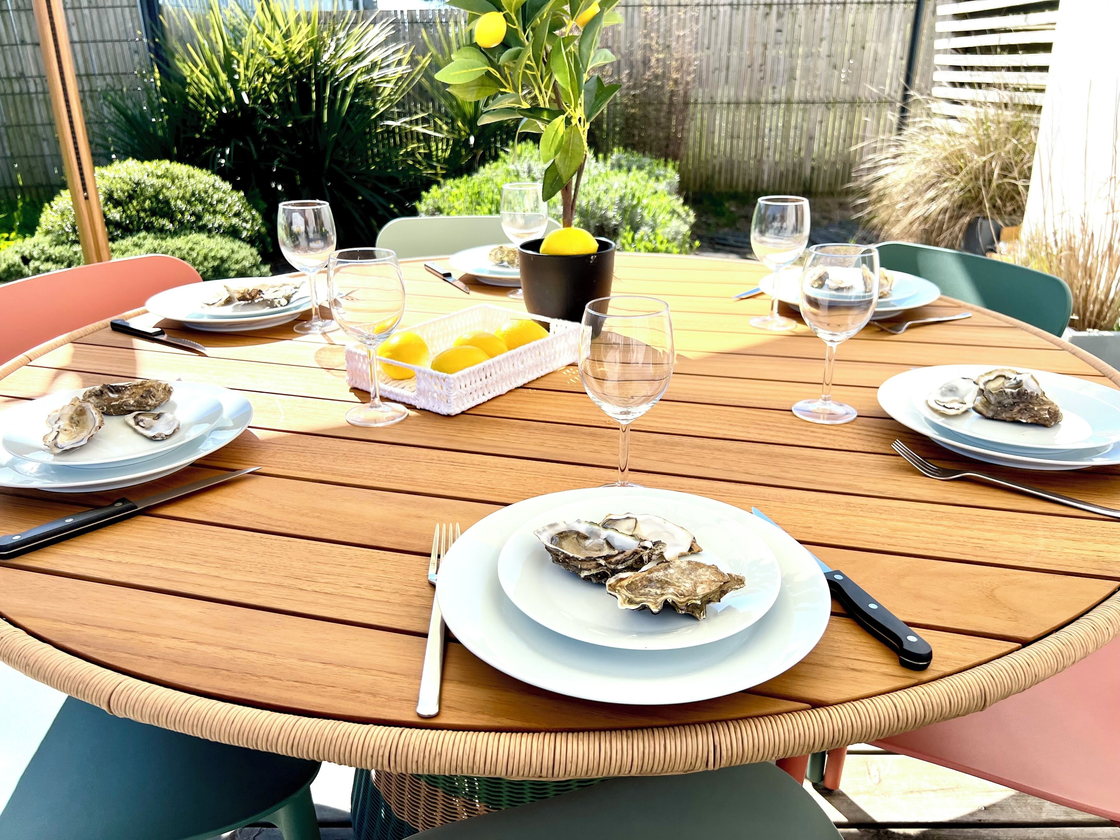 A round wooden table set for a meal with plates of oysters, glasses, and a basket of lemons.