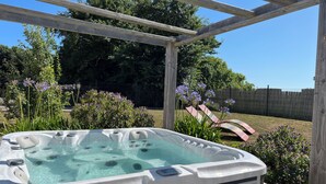 A hot tub in a covered outdoor area with a wooden pergola, surrounded by lush greenery and purple flowers. - Koquillage, Cozy House with Spa, GR34, Sleeps 10, Near the Ocean (Moëlan-sur-Mer)