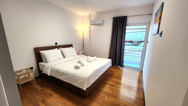 A neatly made bed with white linens, a bedside table with a lamp, and a view of the outside through a window with curtains. - Grec Maison Piraeus by GrecCollection (Piraeus)