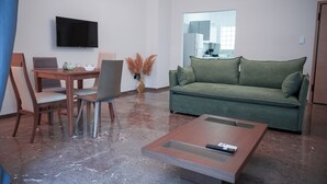 A modern living room with a green sofa, a wooden coffee table, a dining table with chairs, a flat-screen TV, and a vase with pampas grass. - Grec Maison Piraeus by GrecCollection (Piraeus)