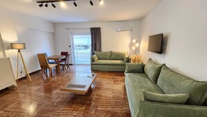 A modern living room with a green sofa, a dining table with chairs, a coffee table, and a TV mounted on the wall. - Grec Maison Piraeus by GrecCollection (Piraeus)
