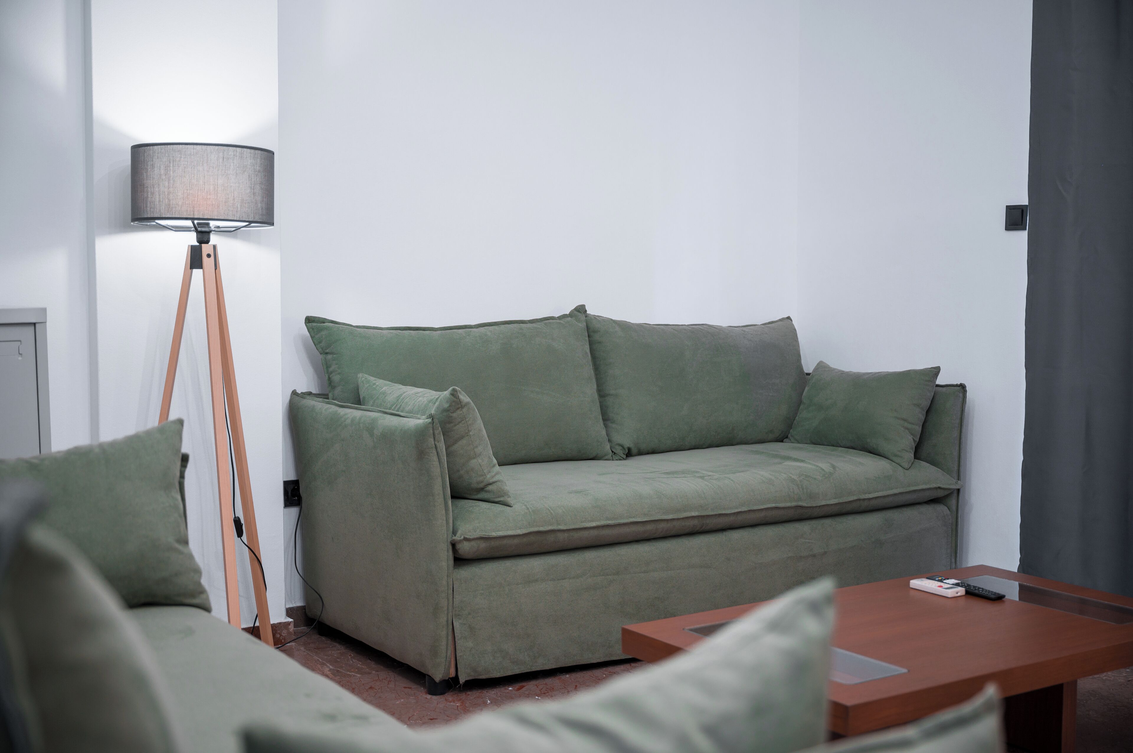 Een groene bank met bijpassende kussens, een houten vloerlamp en een houten bijzettafeltje met een afstandsbediening.