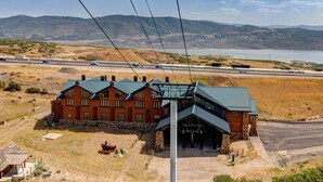 A large log cabin with a metal roof, surrounded by a stone foundation, set against a backdrop of mountains and a highway.