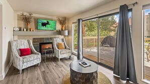 A living room with two patterned armchairs, a fireplace, a television mounted on the wall, and a large sliding glass door leading to an outdoor deck.