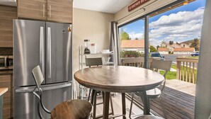 A modern kitchen with a stainless steel refrigerator, a wooden dining table, and a view of a residential area through large windows.