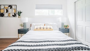 Ein Schlafzimmer mit einem Bett, zwei Nachttischen, einem Fenster mit Jalousien, einem Wandbild und einer Tür.
