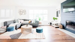 Ein modernes Wohnzimmer mit einem grauen Sofa, runden Korbstühlen-Poufs, einem weißen Couchtisch und einem Kamin.