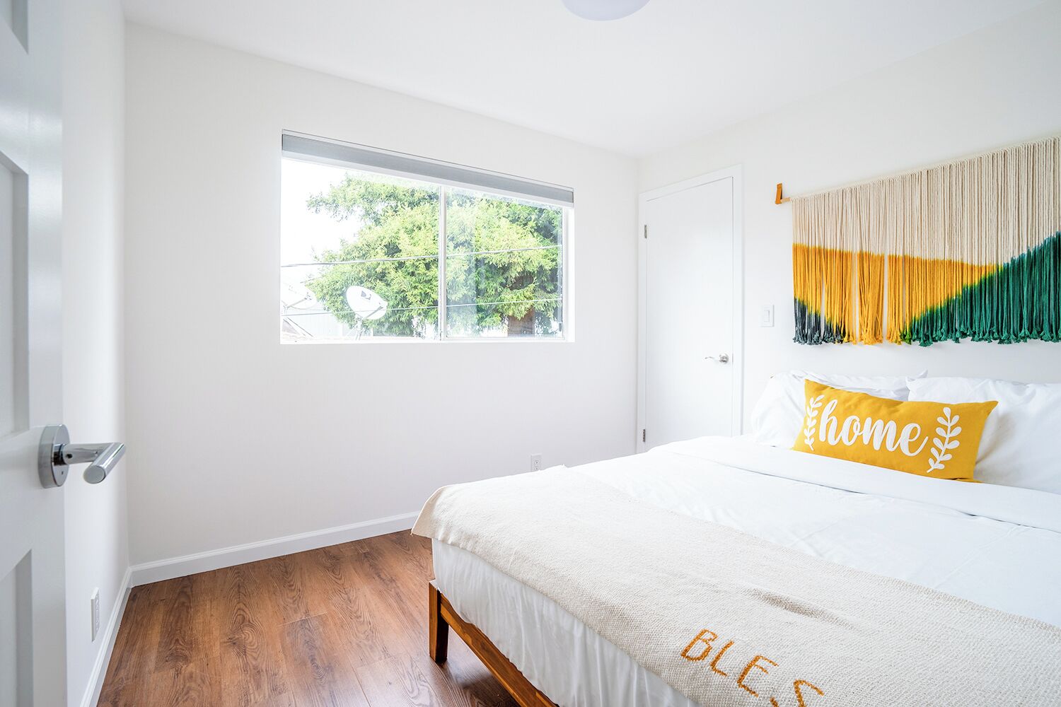 A bedroom with a large window, a bed with white bedding, and a wall-mounted macrame piece.