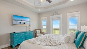 A bedroom with a large bed, a ceiling fan, a TV on the wall, and a view of the outdoors through the windows. - B3 Sand Ranch: 4  BR, 3  BA House in Crystal Beach, Sleeps 14 (Crystal Beach)