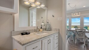 A bathroom with a large mirror, two sinks, and a vanity with drawers. There is a chandelier above the mirror and a hanging light fixture in the dining area. - B3 Sand Ranch: 4  BR, 3  BA House in Crystal Beach, Sleeps 14 (Crystal Beach)