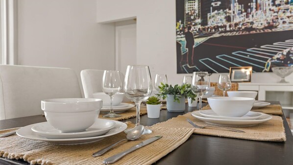A beautifully set dining table with white dinnerware, cutlery, and a woven placemat, surrounded by potted plants and a framed picture.
