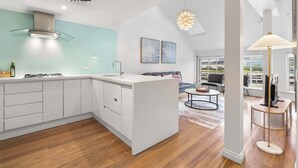 A modern kitchen with white cabinetry, a central island, and a dining area with a round table. - Loft-Style Waterfront Apartment (Woolloomooloo)