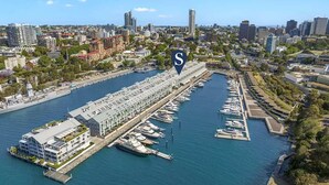 A marina with multiple boats docked, a large building with a sloped roof, and a cityscape in the background. - Loft-Style Waterfront Apartment (Woolloomooloo)