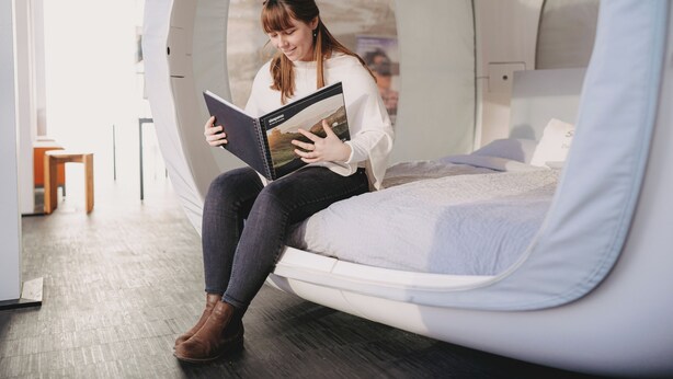 A person sitting on a bed in a room with a window, reading a book. - Eine Nacht im Museum - LUNA Sleep Cube (Lueneburg)