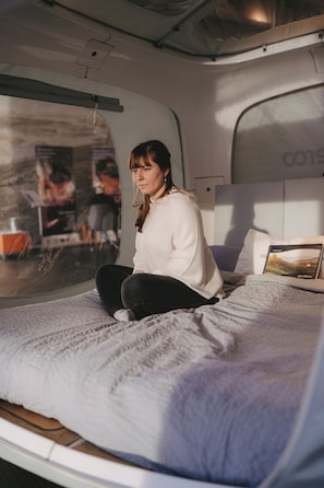Eine Person sitzt auf einem Bett in einem Fahrzeug, das ein Fenster hat. Es gibt ein Kissen und ein gerahmtes Bild auf dem Bett.