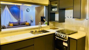 A modern kitchen with a built-in oven, a sink, and a large mirror reflecting a living room with a sofa and curtains. - Jac-Kuw Splendour Homes (Accra)