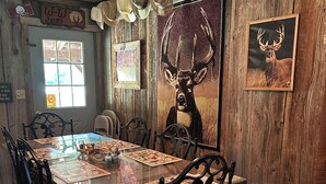 A rustic dining area with a wooden table, black metal chairs, and a deer painting on the wall. - Charming 3-bedroom cabin in Autaugaville with AC and Hangout Building (Autaugaville)