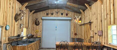 A rustic cabin interior with wooden walls, a dining area with metal chairs, a leather couch, and mounted animal heads.