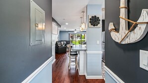 Un pasillo con una pared gris oscuro, una puerta blanca y una pared azul claro con un ancla decorativa. Hay un sofá de cuero negro y una zona de bar con taburetes.