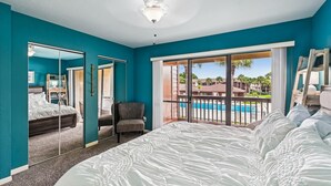 Un dormitorio con una cama grande, una silla, un ventilador de techo y vistas a una piscina y edificios.