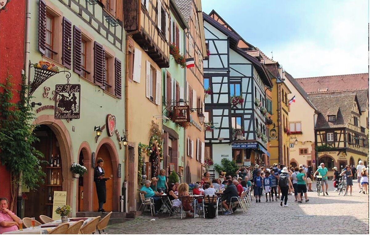 Une rue pavée bordée de bâtiments européens traditionnels, avec des terrasses et une foule de personnes.