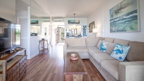 A spacious living room with a large flat-screen TV, a beige sofa with decorative pillows, a wooden coffee table, and a ceiling fan. - Menehune Shores 601 2 Bedroom Condo (Kihei)