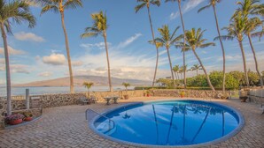 A swimming pool with a stone border, surrounded by palm trees and a stone wall, with a view of the ocean and hills in the background. - Menehune Shores 601 2 Bedroom Condo (Kihei)