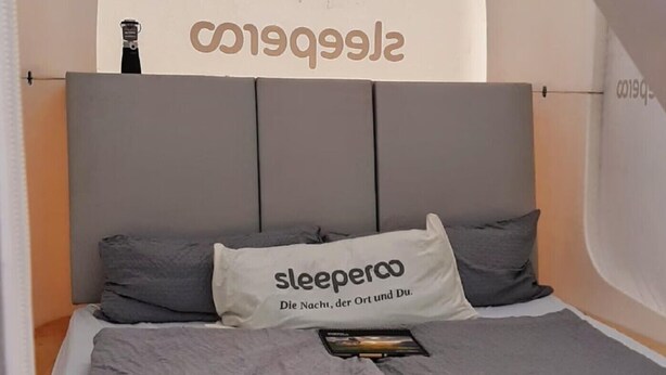 Una cama con cabecera gris, un almohadón con el texto "sleeperoo Die Nacht, der Ort und Du" y un libro sobre la cama.