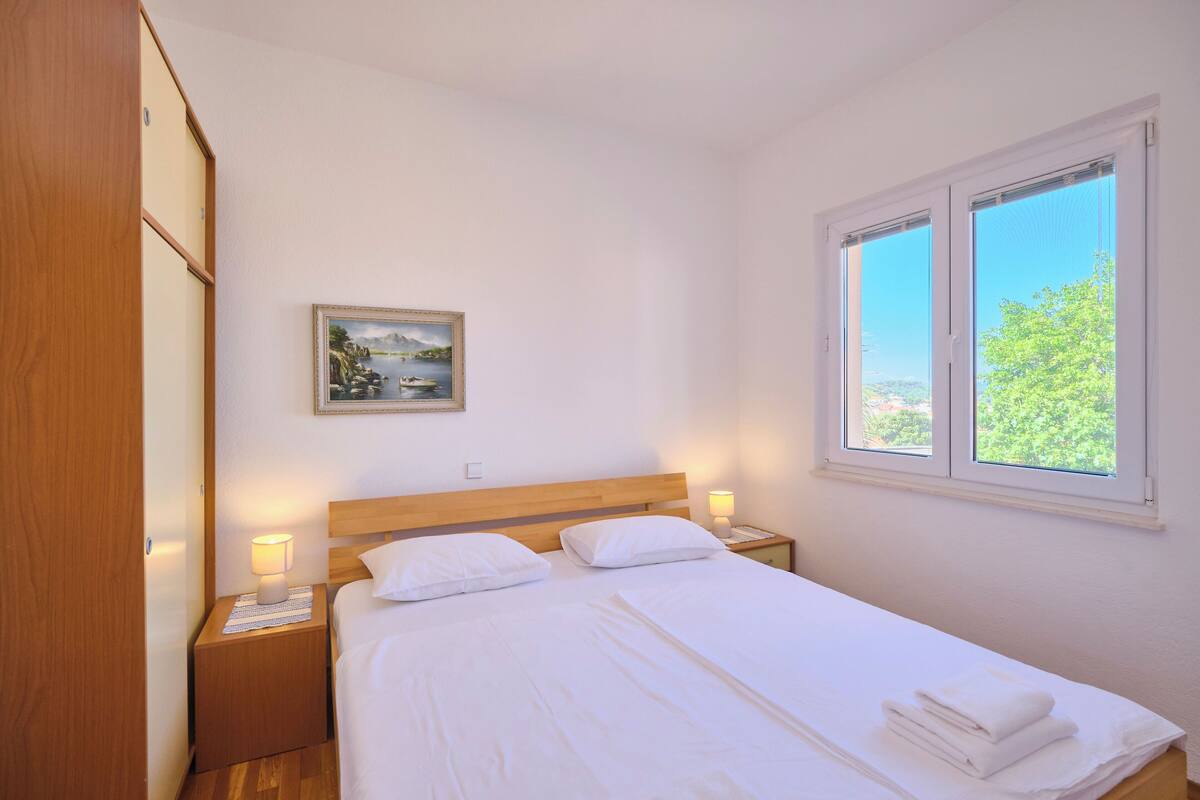 A neatly made bed with white linens and pillows, a bedside lamp, a framed picture on the wall, and a window with a view of greenery.