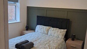 Ein Schlafzimmer mit einem Bett, einem Nachttisch, einer Lampe und einem Fenster.