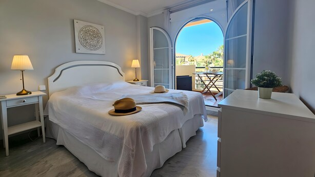 A bedroom with a bed, bedside lamps, a bedside table, a potted plant, and a view through an open door. - Aloha Banús Sea Views by Lica Homes (Marbella)