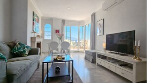A modern living room with a sofa, a coffee table, a TV, and a dining area with chairs and a table. - Aloha Banús Sea Views by Lica Homes (Marbella)