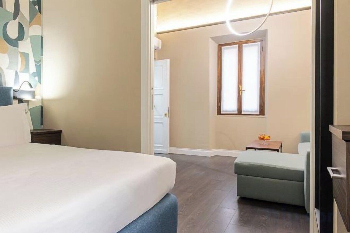 Une chambre d’hôtel comprenant un lit, un petit coin salon, une fenêtre et une porte donnant sur une autre pièce.