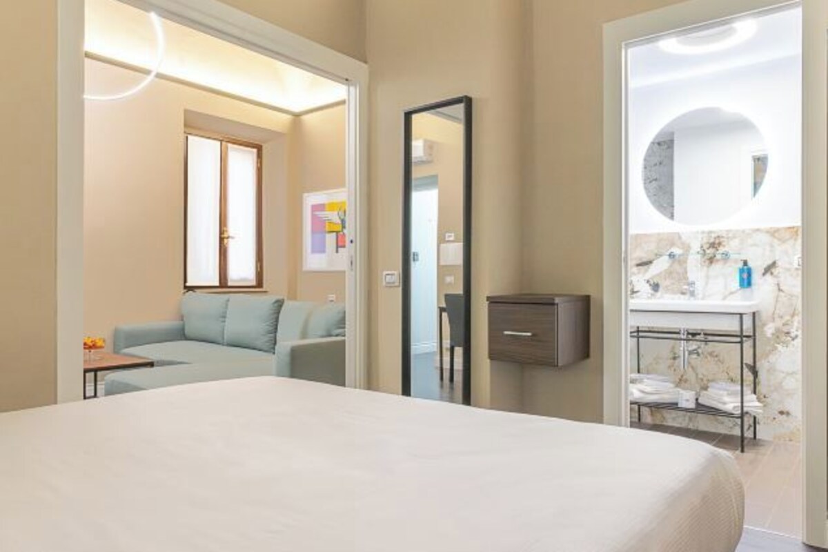 Habitación de hotel con cama, sofá, espejo y baño con lavabo y toallero.