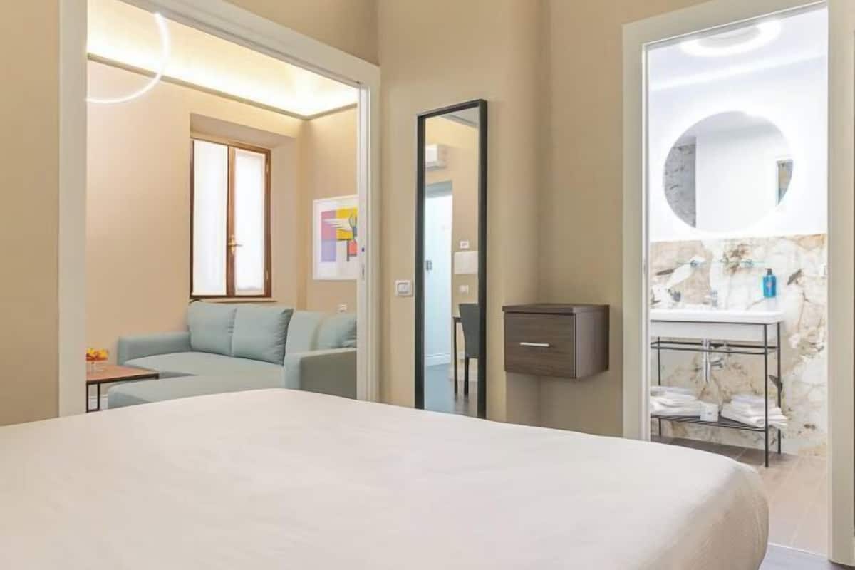 Une chambre d’hôtel comprenant un lit, un canapé, un miroir et une salle de bain avec un lavabo et un porte-serviettes.