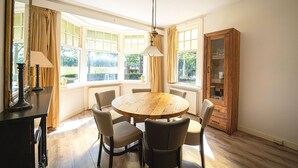 A dining area with a round wooden table, four chairs, a cabinet with glass doors, and large windows with curtains. - Rural Home Near the Soesterduinen (Soest)