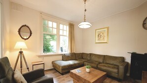 A living room with a sofa, a coffee table, a lamp, a radiator, and a window with curtains. - Rural Home Near the Soesterduinen (Soest)