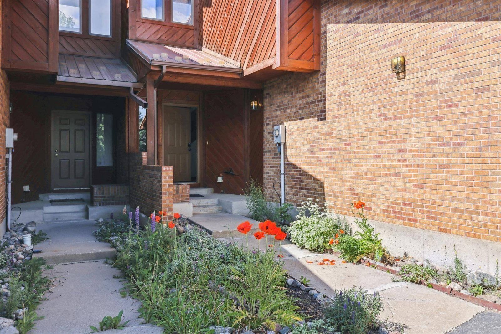 Ein zweistöckiges Haus mit Ziegelaußenwand, einer Holzveranda und einem kleinen Garten mit roten Blumen.