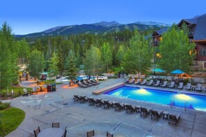 A pool area with lounge chairs and a pool, surrounded by trees and mountains in the background. - Grand Timber Lodge 2 Bedroom Ski In, Ski Out Condo (Breckenridge)