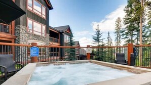 Un bain à remous bordé de pierres, avec une terrasse en bois et une vue sur un chalet doté d’une façade en pierre et de grandes fenêtres.