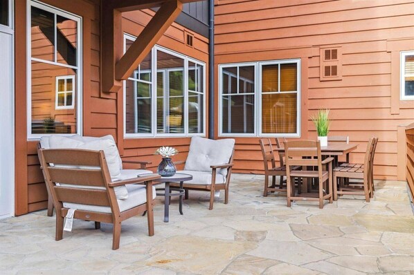 Un patio exterior con mobiliario de madera, suelo de piedra y ventanas amplias.