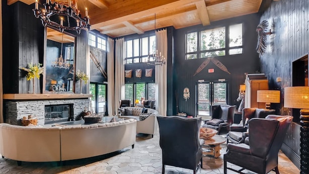 A spacious living room with a stone fireplace, leather sofas, and a large window overlooking a forest. - Bluesky Breckenridge Luxury 3 Bedroom Condo at Snowflake Lift (Breckenridge)