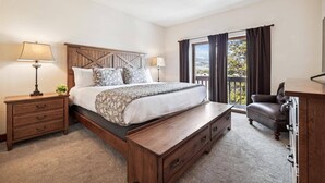 Une chambre avec un grand lit, une tête de lit en bois, deux tables de chevet avec des lampes, un banc en bois, un fauteuil en cuir et une vue sur l’extérieur par une fenêtre.