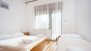Una habitación de hotel con dos camas, piso de madera, un ventanal grande con cortinas translúcidas y una mesita de noche con lámpara.