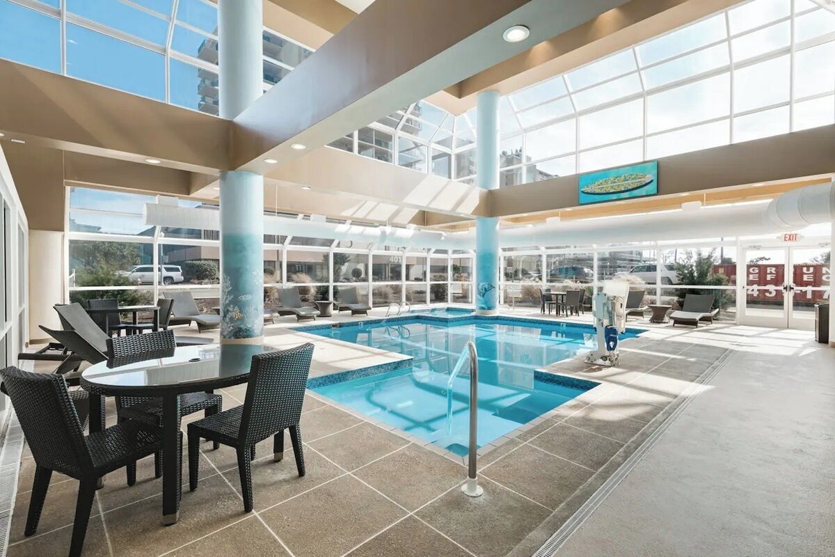An indoor swimming pool with a glass ceiling, surrounded by lounge chairs and a dining area with a table and chairs.