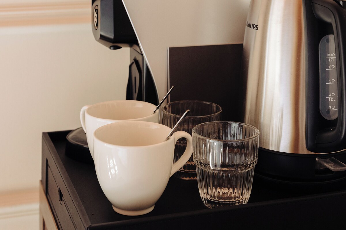 A coffee maker, two white cups, and a glass on a black tray.