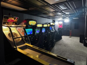 Uma fileira de máquinas de jogos de fliperama em um espaço com pouca luz, chão coberto de carpete e uma porta ao fundo.